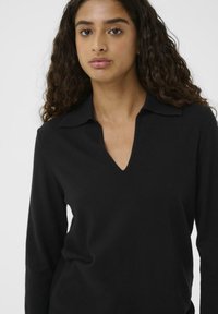 Giovane donna con capelli ricci che indossa un maglione nero a maniche lunghe con scollatura a V e colletto, su uno sfondo chiaro e semplice.