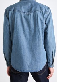 Camicia di jeans blu chiaro con maniche lunghe, polsini a bottone e orlo curvo. Presenta un'ampia spalla con un sottile design a forma di V.