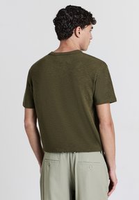 Uomo con capelli ricci che indossa una camicia a maniche corte verde oliva e pantaloni grigio chiaro, rivolto di spalle su uno sfondo bianco uniforme.