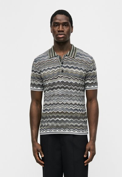 Άνδρας που φοράει κοντομάνικο πλεκτό πόλο με γιακά, με μαύρα, λευκά και χακί ζιγκ-ζαγκ μοτίβα και μαύρο παντελόνι.