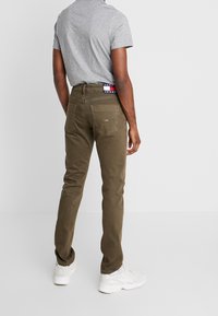 Vaqueros ajustados color verde oliva con cinco bolsillos, tejido vaquero, detalles de costuras y cinturilla con logotipo; combinados con una camiseta gris.