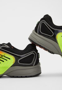 Paire de chaussures de sport noires et grises avec des accents jaune néon et orange, montrant les vues latérales et arrière sur fond blanc.