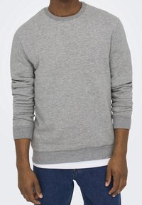 Szary sweatshirt z okrągłym dekoltem, prążkowanymi mankietami i dołem. Miękka tekstura z delikatnym melanżowym wzorem, założony na białą koszulę.