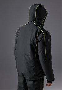 Chaqueta negra aislante con costuras amarillas, capucha y un logo en la manga. Presenta un corte recto y una tela suave y resistente al agua.