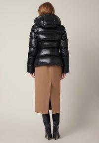 Czarna kurtka puchowa z kapturem, o błyszczącej powierzchni i poziomym pikowanym wzorze. Nosi ją nad brązową spódnicą i czarnymi botkami do kolan.