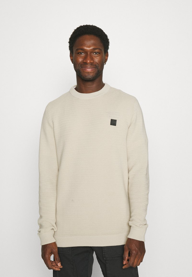 Vīrietis ar īsiem cirtainiem matiem, valkājot krēmkrāsas ribotu garroku džemperi un melnas bikses, smaida uz vienkārša balta fona.