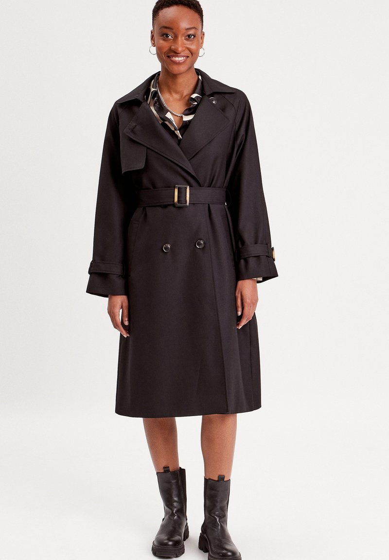 Sort trenchcoat med dobbeltknap design, bælte i taljen, brede revers og knapmansjetter. Båret over en mønstret skjorte, parret med ankelstøvler.