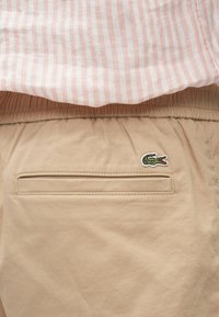 Pantalon khaki avec ceinture élastique, poche arrière unique et logo de crocodile vert brodé. Tissu texturé avec une finition lisse.