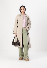 Beiger Trenchcoat mit dunklen Knöpfen, hellrosa Oberteil, grüne, weit geschnittene Hose und eine kleine braune Schultertasche. Gestylt mit beigen Schuhen.