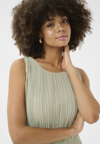 Ljust grön plisserad klänning med rund halsringning. Tyget har en texturerad, flödande yta som kompletteras av modellens lockiga hår.