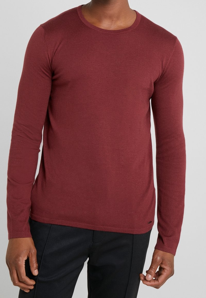 Pull col ras du cou à manches longues couleur bordeaux en tissu doux, présentant une coupe ajustée et des poignets côtelés, assorti à un pantalon foncé.