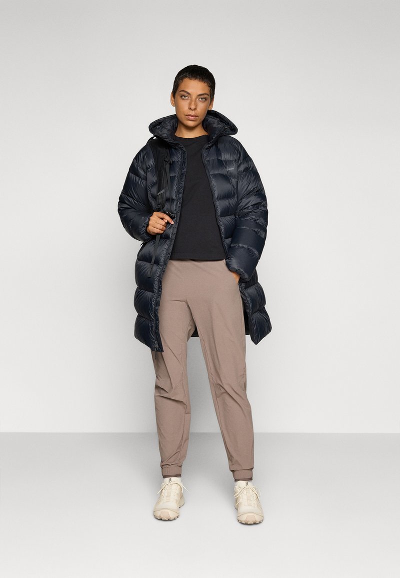 Marineblå pufferjakke med høj krave og glansfuld overflade, parret med beige tapered bukser og lyse sneakers. Enkel design uden mønstre.