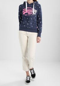 Felpa blu navy con cordini azzurri, grafiche stampate in rosa e bianco, abbinata a pantaloni bianchi a vita alta e sneakers nere.