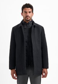 Zwarte wollen jas gelaagd over een zwarte pufferjack, met een geknipte kraag, knoopsluiting en gestructureerde stof. Grijze broek zichtbaar.