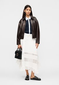 Žena v tmavohnedej koženej bunde, námornícky modrom zapínanom topu, bielej sukni zdobenej čipkou, čiernych mokasínach, držiaca čiernu kabelku.