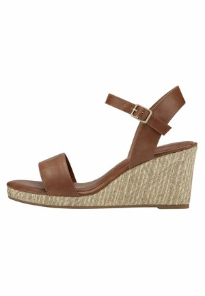 Brązowe skórzane sandały na koturnie z szerokim paskiem na przodzie i paskiem wokół kostki z klamrą, osadzone na teksturowanej, plecionej podstawie.