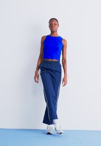 Kék ujjatlan cropped top, sötétkék, elasztikus derekú nadrág fehér oldalcsíkokkal, és ezüst színű sportcipő. Egyszerű, világos háttér.