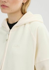 Lichtcrème zip-up hoodie met een zachte textuur en een voorzijde met een reliëflogo. Voorzien van een trekkoordcapuchon en raglanmouwen, wat zorgt voor een ontspannen pasvorm.