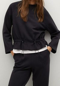 Sudadera negra con cintura ajustable, cuello redondo y mangas largas, combinada con pantalones negros a juego. Textura de tela suave.