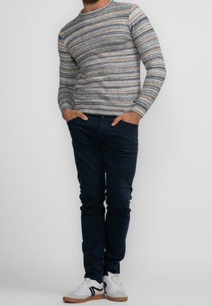 Hombre con suéter de rayas de manga larga, pantalones oscuros ajustados y zapatillas blancas, de pie con las manos en los bolsillos frente a un fondo liso.