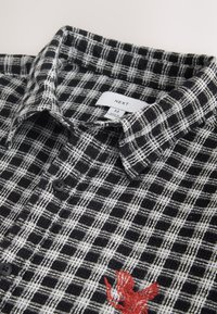Chemise à carreaux noir et blanc à boutons avec un oiseau rouge brodé en bas à droite et un col plié.