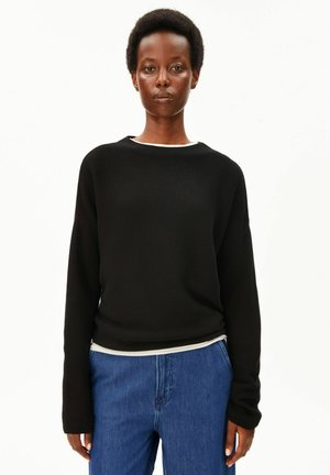 Femme aux cheveux courts et bouclés portant un pull noir à manches longues par-dessus une chemise blanche et un jean bleu, debout devant un fond blanc uni.