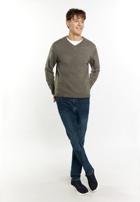 Maglione grigio a maglia con scollo a V, abbinato a jeans blu e sneakers scure. Modello in piedi con una postura rilassata su uno sfondo bianco.