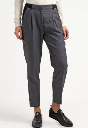 Femme portant un pantalon gris sur mesure longueur cheville avec plis, passants de ceinture noirs et chaussures slip-on en cuir noir avec semelles épaisses.