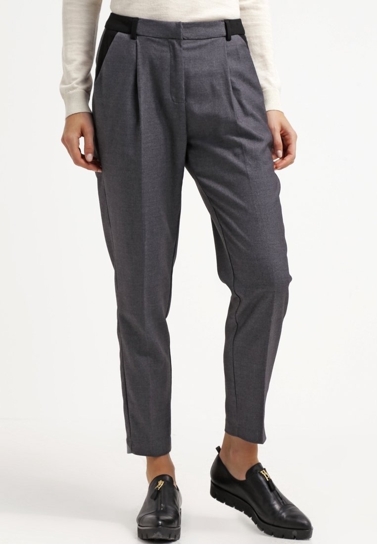 Femme portant un pantalon gris sur mesure longueur cheville avec plis, passants de ceinture noirs et chaussures slip-on en cuir noir avec semelles épaisses.
