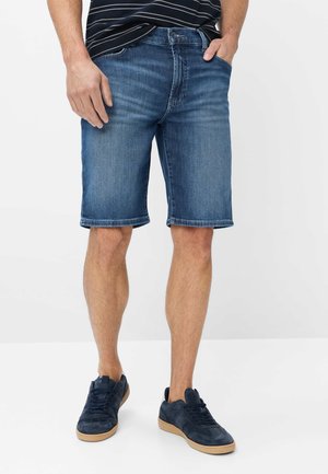 Mand iført mellemblå denimshorts, marineblå sneakers med brune såler og en sort stribet skjorte med hænderne i lommerne.