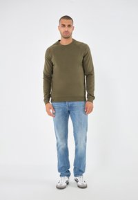 Sudadera verde oliva de mangas largas y cuello redondo, combinada con jeans azul claro y zapatillas blancas con acentos negros.