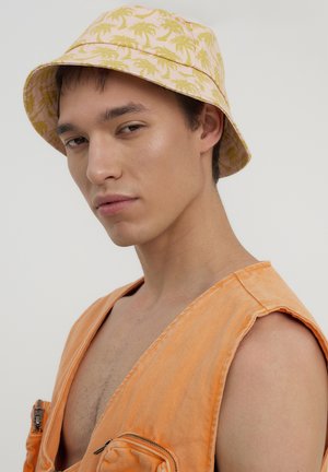 Cappello a secchiello realizzato in tessuto rosa con motivi di palme gialle. Indossato con un indumento senza maniche arancione e una finitura testurizzata.