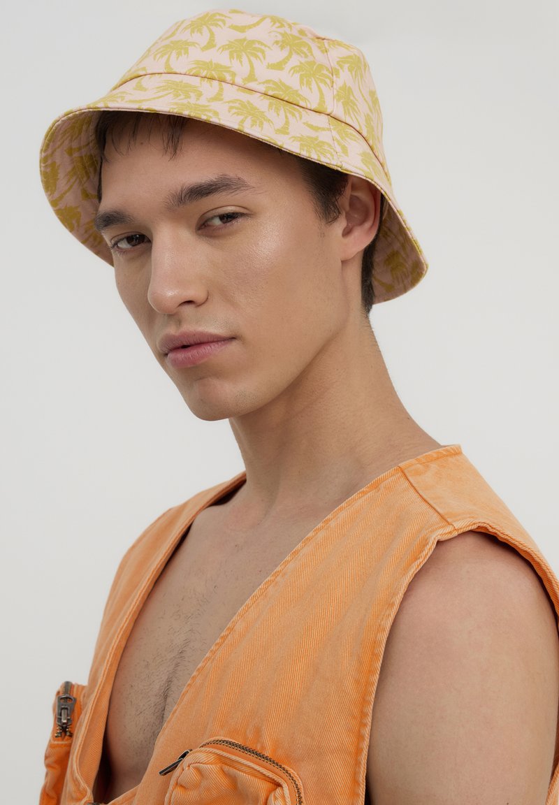 Cappello a secchiello realizzato in tessuto rosa con motivi di palme gialle. Indossato con un indumento senza maniche arancione e una finitura testurizzata.