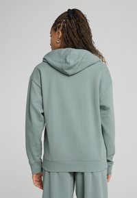 Mikina z bavlněné směsi v tlumené zelené barvě, s velkou kapucí, volným střihem, žebrovanými manžetami a lemem; hladká textura a minimalistický design.