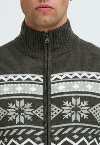 Mörkgrön zip-up tröja med hög krage. Har vita och ljusgröna snöflinge- och geometriska mönster över bröstet.