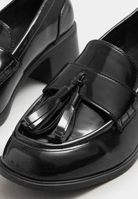 Svarta lackskinnsloafers med en glänsande yta, utrustade med en dekorativ tofs och en kraftig klack för extra höjd.
