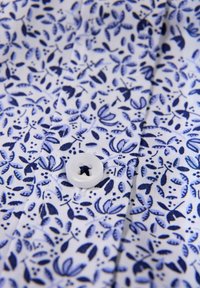 Witte stoffen blouse met een blauw bloemmotief, met een rond witte knop deels zichtbaar en een gladde textuur.