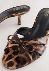 Mules à talons ouverts à imprimé léopard avec accents noirs, ornées d'un nœud noué et d'une texture lisse et brillante.