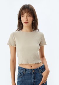 Beige ribbad crop top med rund halsringning och korta ärmar. Scalloped fålldetalj. Bärs med högmidjade blå denimjeans.