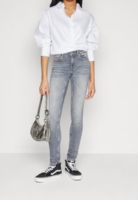 Camisa blanca de botones, jeans ajustados de un azul claro, zapatillas negras con suelas blancas y un bolso plateado. Tejido suave y diseño ajustado.
