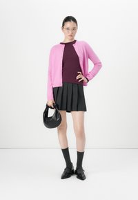 Vrouw met een bril, roze cardigan over een bordeaux top, zwarte geplooide rok, donkere sokken met zwarte schoenen, die een zwarte handtas vasthoudt tegen een witte achtergrond.