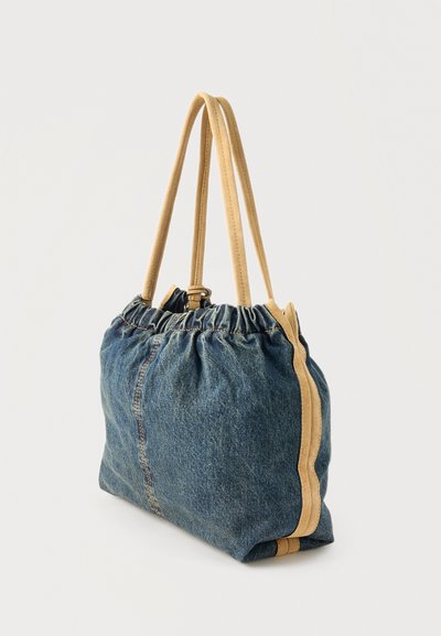 Μαλακή τσάντα tote από τζιν με δερμάτινα χερούλια και επένδυση σε ταμπά χρώμα, με συγκεντρωμένη πάνω άκρη, σε ορθια θέση μπροστά σε απλό ανοιχτό φόντο.