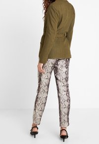 Olivově zelený sako s páskem a texturovaným povrchem, spárované s přiléhavými kalhotami se vzorem hada v béžových a hnědých tónech. Stiletto podpatky.