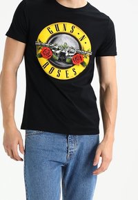 Zwarte katoenen T-shirt met een grafisch ontwerp met "GUNS N' ROSES," rozen en een zilveren gitaar, ronde logo-stijl druk. Korte mouwen.
