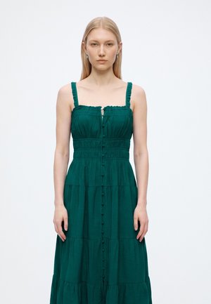 Jeune femme aux cheveux blonds raides, portant une robe verte sans manches boutonnée, debout devant un fond blanc uni.