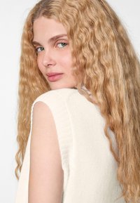 Helles, ärmelloses Oberteil mit geripptem Textur, weichem Stoff und rundem Ausschnitt, getragen von einem Model mit langen, gewellten blonden Haaren.