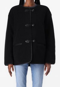 Chaqueta de forro polar negra con cuello redondo, cierres de botones y bolsillos laterales. El diseño presenta una textura suave y un ajuste relajado y holgado.