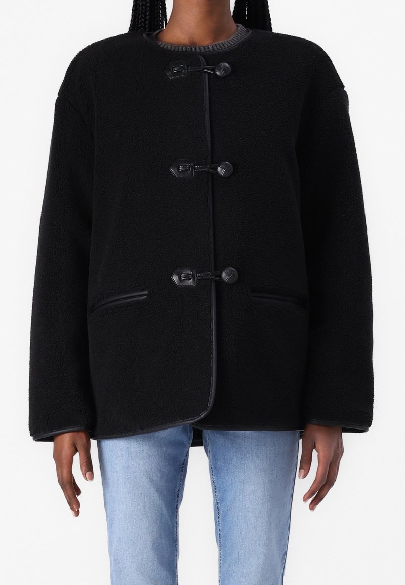 Chaqueta de forro polar negra con cuello redondo, cierres de botones y bolsillos laterales. El diseño presenta una textura suave y un ajuste relajado y holgado.