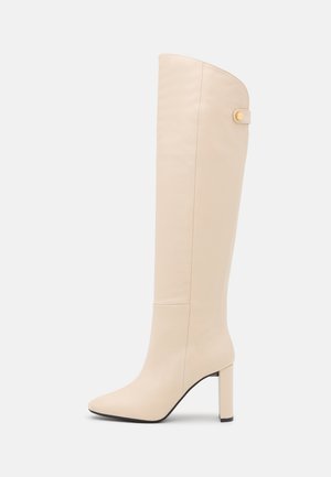 Roomkleurige leren laars tot aan de knie met blokhak en een klein riempje met gouden knopje bij de bovenrand.