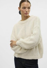 Pull à la coupe oversize de couleur crème avec une texture en tricot, un col côtelé et des manches ballon. Associé à un jean noir. Design minimaliste.